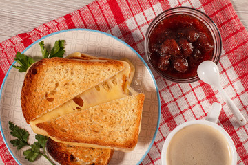 Toasted slices of bread with cheese and green parsley on white plate, cup of coffee, bowl with strawberry jam and spoon on napkin.