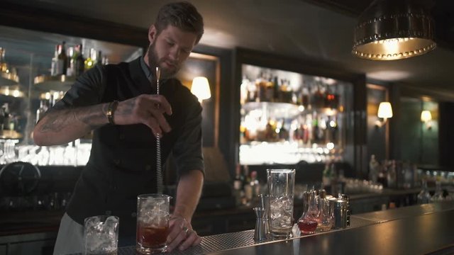 Attractive bearded bartender mixing rum and ice in the glass with long metal stick. Barman making cocktail in modern bar with many bottles on shelves