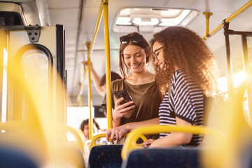Two girls watching phone and smiling while standing on a bus. - Powered by Adobe