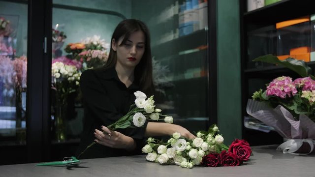 Professional woman florist cuts white and red roses with a pruner over the table in flower shop.
