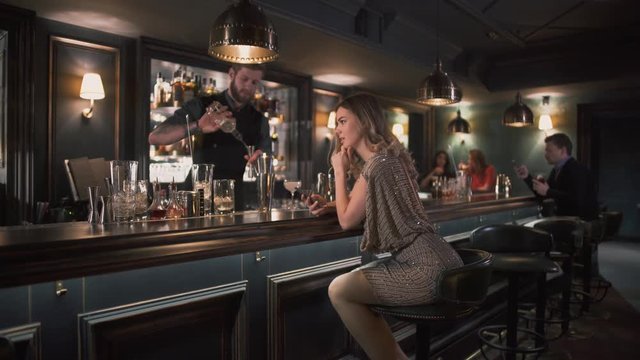 Hipster bartender combining ingredients and making cocktails in bar while young pretty woman sits near bar counter and exting message