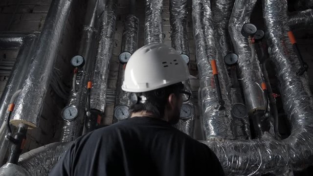 Man in white helmet stands with his back to camera opposite the pipe with pressure sensors in boiler room. He conducts an inspection of pipes, writes indicators. Checking heating, ventilation system