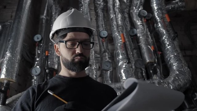 Man with beard, wearing glasses and white helmet stands in a boiler room, inspects. Against the background of hot pipes in a silver corrugation, pressure sensors. He takes notes, looks at the sensor.