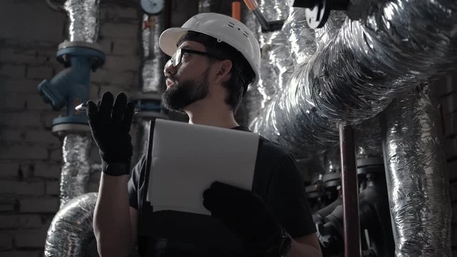 Male engineer works in boiler room and dictates audio message on smartphone. Plumber in technical room speaks in the speaker phone (speakerphone, audio message or voice recorder). Pipes on background