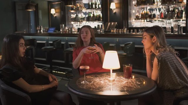 Three young girlfriends in evening dresses sitting at the small table in bar close up. Girls have celebration together, they chatting and smiling. Lonely ladies have a fun