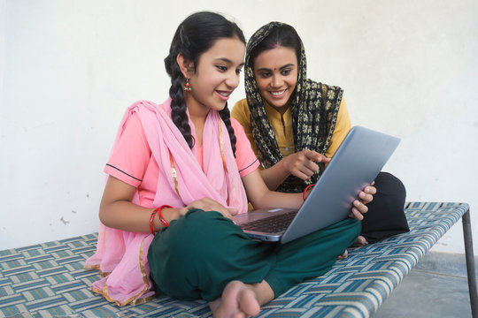 Happy village girl sitting with her mother on a cot operating a laptop computer.	