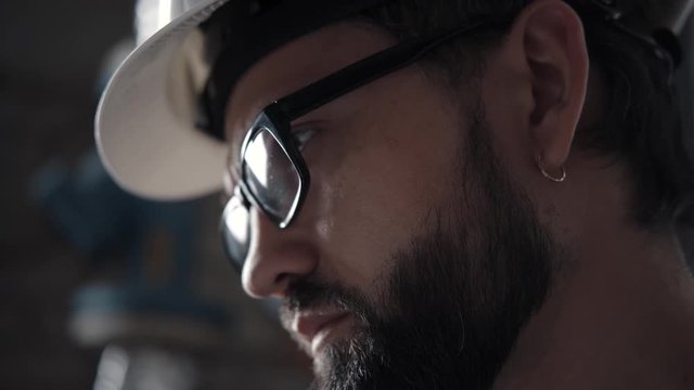 Profile of a man in a helmet, glasses and a beard: he looks down at the sheet of notes. A technical specialist (engineer) works in a boiler room, industrial room. He has a earring in his ear.