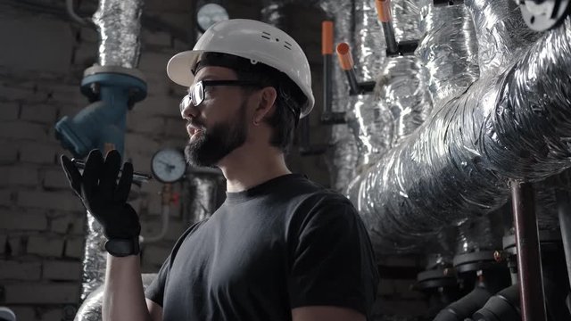 Male engineer works in boiler room and dictates audio message on smartphone. Plumber in technical room speaks in the speaker phone (speakerphone, audio message or voice recorder). Pipes on background