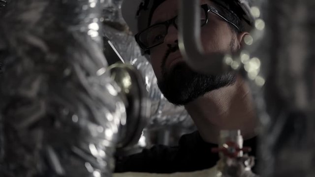 Man in helmet and glasses works in room with pipes (heating or gas station), carefully studies sensors, checks the system. Safety. Portrait is shot through the pipes in a silver corrugation.