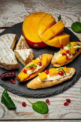 Bruschetta with mango, Camembert cheese and pomegranate on a old rustic table. Traditional italian appetizer or snack, antipasto served on a shale board. Top view