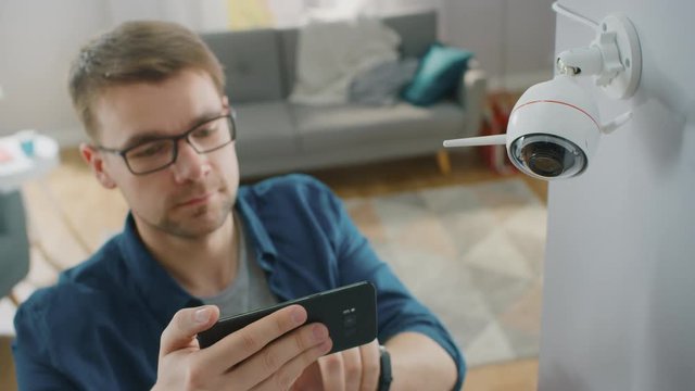 Young Man in Glasses Wearing a Blue Shirt is Adjusting a Modern Wi-Fi Surveillance Camera with Two Antennas on a White Wall at Home. He's Checking the Video Feed on his Smartphone.