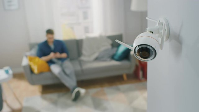 Close Up Object Shot of a Modern Wi-Fi Surveillance Camera with Two Antennas on a White Wall in a Cozy Apartment. Man is Sitting on a Sofa in the Background.