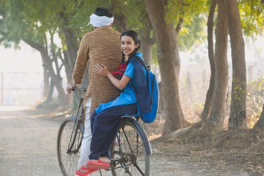 Rear view of a happy school going girl pillion riding on bicycle with her father in village.	