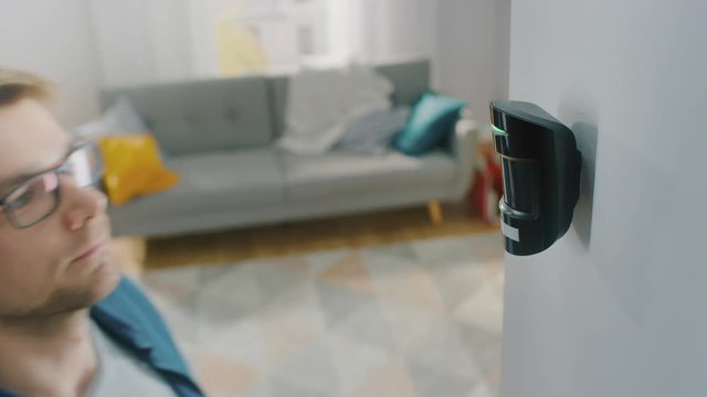 Young Man in Glasses Wearing a Blue Shirt is Hanging a Modern Movement Detector Unit on a White Wall at Home. Device Blinks with Green Led Light.