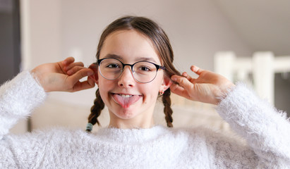 Portrait of funny attractive smiling preteen girl in glasses with pigtails making faces bulging ears and sticking her tongue out fooling around. Having fun at April fools day. Playful childhood