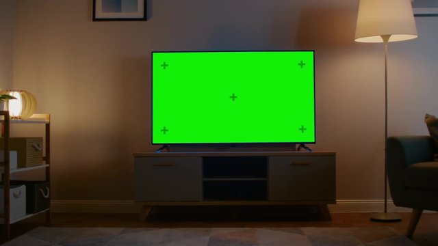 Zoom Out Shot of a TV with Horizontal Green Screen Mock Up. Cozy Evening Living Room with a Chair and Lamps Turned On at Home.