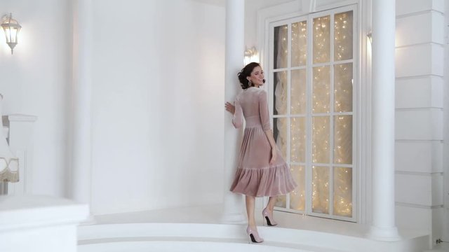 elegant woman in dress posing next to a high column and a large window, walking up the stairs, slow motion