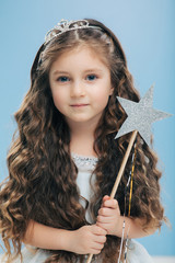 Small kid angel has long crisp hair, blue eyes, holds magic wand in form of star, poses over blue background, has appealing appearance. Little princess waits for miracle, believes in fairy tale
