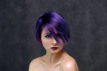 A beautiful, sexy girl with purple hair and a short haircut sits in the middle of the photo with a gray background