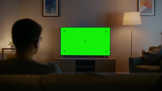 Young Man in Glasses is Sitting on a Sofa and Watching TV with Horizontal Green Screen Mock Up. It's Evening and Room at Home Has Working Lamps.