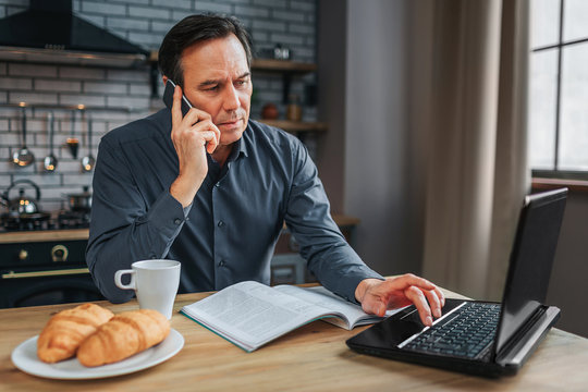 Busy Man Sit At Table In Kitchen And Talk On Phone. He Type On Keyboard Laptop And Work. Journal White Cup And Plate With Croissans On Table.