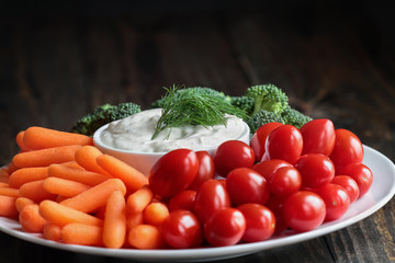 Homemade buttermilk ranch salad dressing with dill served with fresh vegetables, cherry tomatoes, baby carrots and broccoli, over a rustic wooden background.