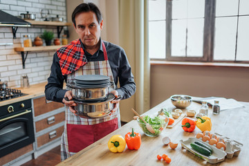 Serious concentrated man in apron stand alone in kitchen. He hold pans and dishes. Guy stand at...