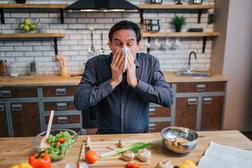 Sick adult man sneezing to white napkin. He stand at table in kitchen. Desk full of colorful healthy vegetables and spices.