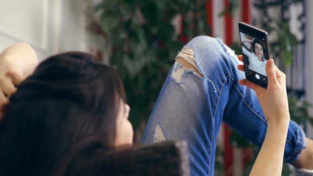 young woman, girl, brunette, in white shirt and jeans, lying on a sofa, small couch, with smartphone. listening to music, looks at screen, takes a selfie video, grimaces. blurred background