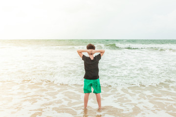 Boy in green shorts and black t-shirt standing by the ocean on the beach, looking at the waves and enjoying current moment
