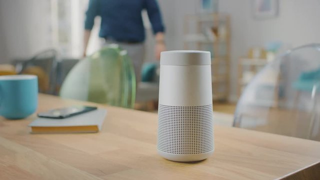 Close Up Shot of a Modern Silver Wireless Speaker Standing on a Table at Home. Man in the Background Stands Up From the Sofa, Walks to the Speaker and Pushes a Button.