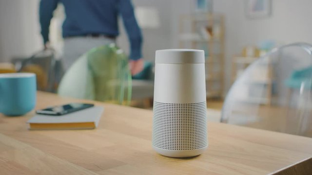 Close Up Shot of a Modern Silver Wireless Speaker Standing on a Table at Home. Man in the Background Walks to the Speaker and Pushes a Button. Then He Sits on a Couch.