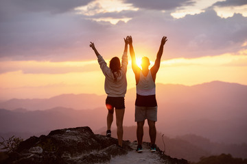 Fototapeta na wymiar Happy couple with raised arms stands on mountain top against sunset and having fun in winner pose