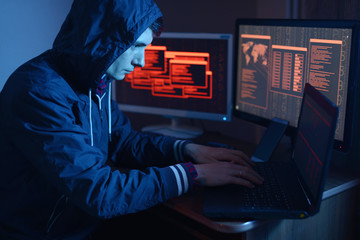 Hacker in a hood typing program code while committing a cybercrime on the background of screens in...