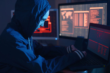 Girl hacker in a hood typing program code while committing a cybercrime hacking a firewall on the background of screens