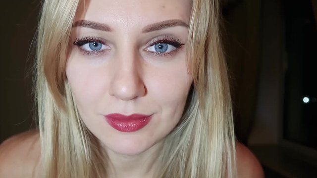 young beautiful blonde woman with bright blue big eyes and red lips flirts at the camera, raises one eyebrow and seduces with her eyes, close-up portrait on a dark background, slow motion