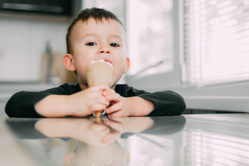 A child in a dark-blue t-shirt in the bright kitchen eating a waffle ice cream cone in the summer house