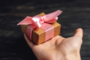 Hand holding Holiday gift box Packed in crafting paper with pink ribbon on dark wooden background