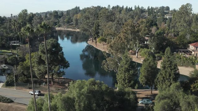 The aerial camera descends from 185 ft. above the ground then flying from the north toward the lake flying over the lake at 120 ft. over the water. Locals walk along the banks of the lake.