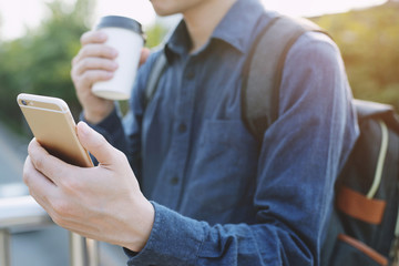 Travel blogger young man using route application using mobile smart phone reading to find the needed address in a city and holding a cup of take away coffee drink in morning sunlight. 