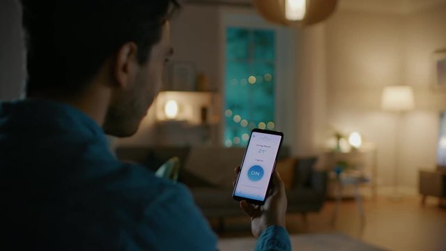 Young Handsome Man Gives a Voice Command to a Smart Home Application on His Smartphone and Lights in the Room are Being Turned On. It's a Cozy Evening.