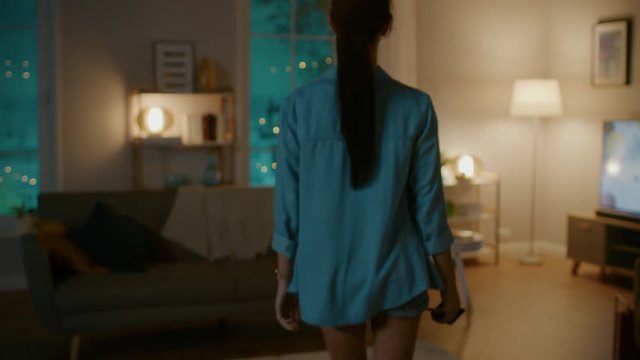 Young Beautiful Woman Gives a Voice Command to a Smart Home Application on Her Smartphone and Lights in the Room are Being Turned On. She Walks and Sits on a Couch. It's a Cozy Evening.
