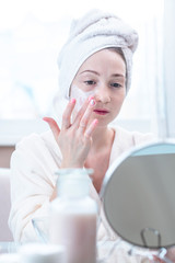 Beautiful happy young woman applying cream on face moisturizing the skin. Hygiene and care for the skin at home