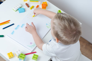 Little child draws in his album on a white table. Pencils, train, colorful plastic block in the children's room.