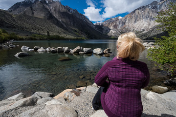 Naklejka premium Blond female sits alone by Convict Lake in the springtime, located off of US-395, near Mammoth Lakes California in the eastern Sierra Nevada mountains, Inyo National Forest.