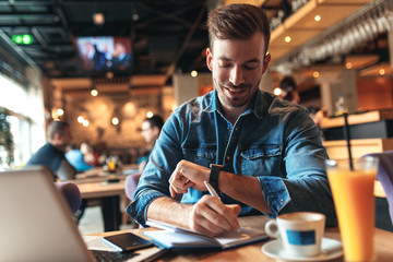 Young man sitting at cafe and working on laptop, casual businessman looking at smart watch.