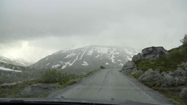 Driving through the Old Strynefjell Mountain Road in Norway with bad weather conditions and mountains with snow in the background in slow motion.
