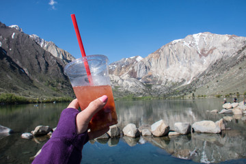 Hand holds a fruity tea drink placed in front of Convict Lake located off of US-395, near Mammoth...
