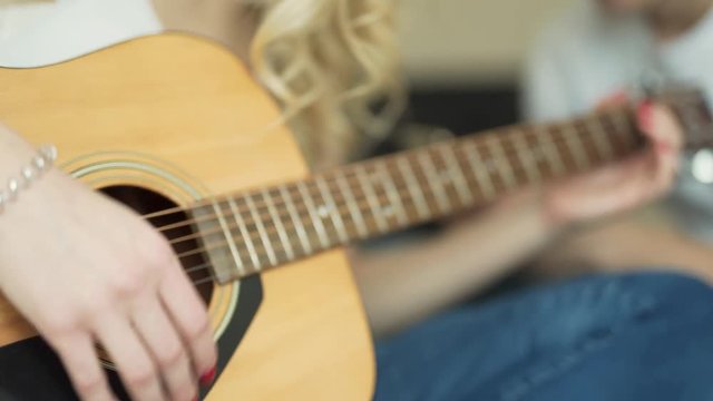 A close up video of a girl who masterly plays on guitar. She tries to use different chords for better sound and melody. The background is blurred. There is a figure of the boy at the background.