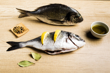 Two fresh fish Dorada with bay leaves, some pieces of lemon, a bowl of oil and some spices on a wooden table